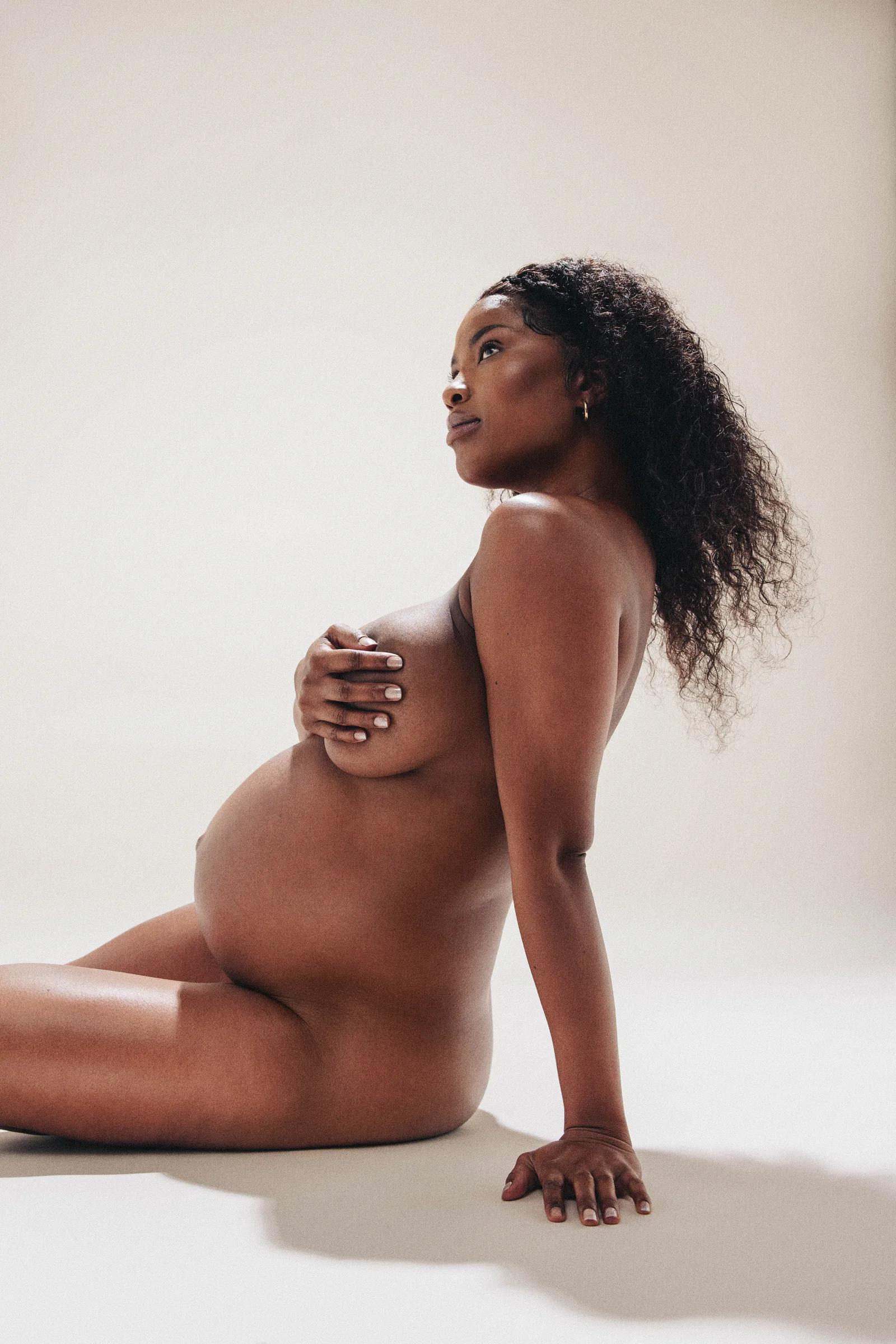 A pregnant woman seated on the floor in soft studio light, one hand on her chest, looking up.